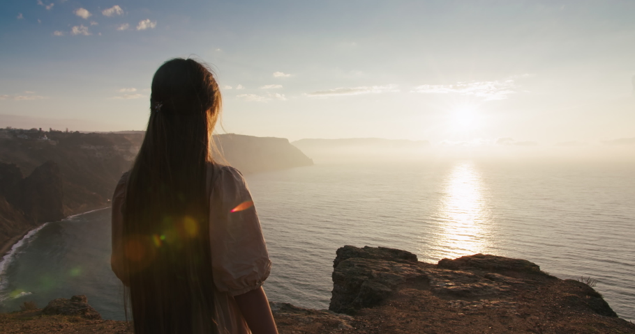 Fairy tale little girl with long hair goes to the edge of cliff to look stunning seascape at early morning. Girl character meeting sunrise on high mountain near sea. Wonderland landscape view Royalty-Free Stock Footage #1076198048