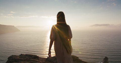 Fairy tale little girl with long hair goes to the edge of cliff to look stunning seascape at early morning. Girl character meeting sunrise on high mountain near sea. Wonderland landscape view