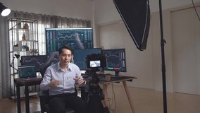 Asian Male Stock Market Broker Teaching How To Trade To Camera With Professional Light Equipment, Analysing Graphs On Multiple Computer Screens At The Background
