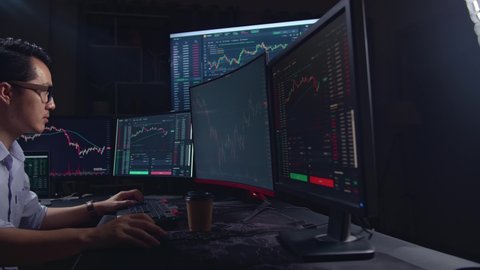 Side View Of A Asian Young Male Stock Market Broker Analysing Graphs On Multiple Computer Screens
