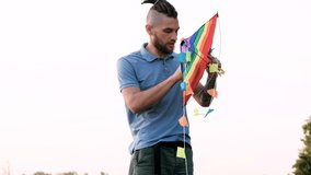 LGBT flag kite. Slow motion 4k video. Young Caucasian man with dreadlocks is standing and trying to untangle kite to launch it and have fun in fresh air.