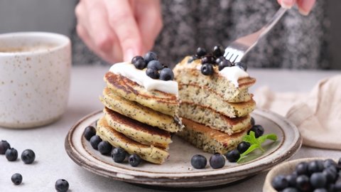 Young woman eating poppy seed pancakes with yogurt and blueberries using fork and knife, cutting stack in halves