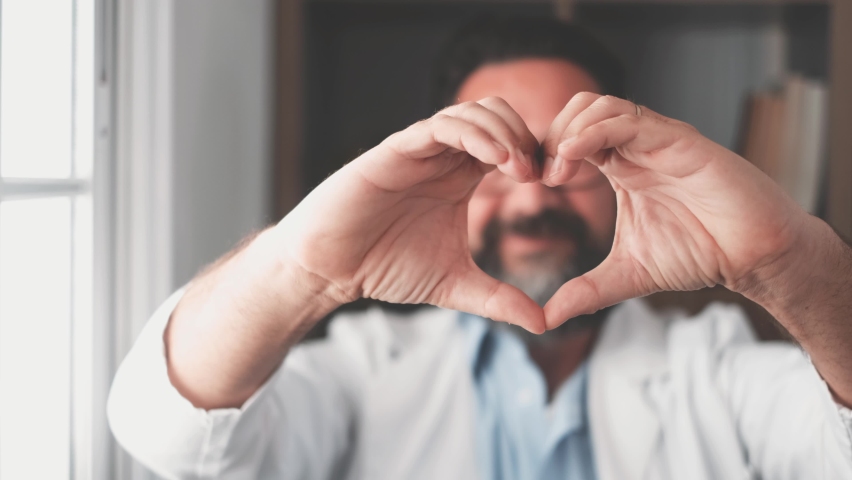 Close up portrait of smiling young caucasian male nurse or GP in white medical uniform show heart love hand gesture. Happy young man doctor show support and care to patients or client in hospital.
 | Shutterstock HD Video #1076203637