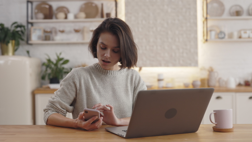 Attractive woman with short haircut using modern smartphone while sitting at kitchen table with portable laptop. Concept of people, technology and distance learning. Royalty-Free Stock Footage #1076204867