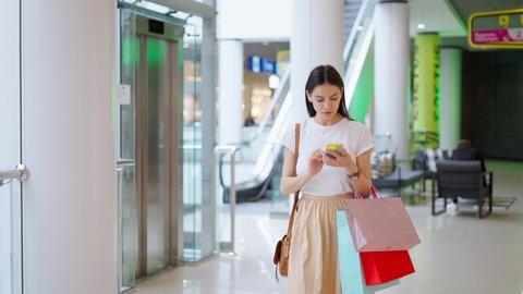 Beautiful young europian woman in stylish outfit with modern smartphone and many paper shopping bags using real time navigation application to find store in mall or commercial center