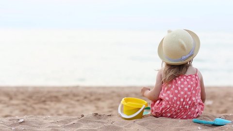 baby girl is sitting on beach and playing in sand in summer. holidays with kids. child turns around, smiles and waves his hand