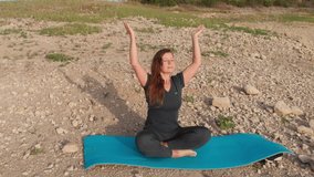 Drone video of a young woman in sportswear sitting in the morning with her legs crossed,arms raised on the shore of a beautiful lake in the yoga lotus exercise pose.Yoga classes,meditation in nature