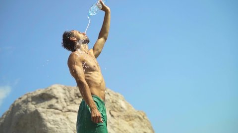 A young guy of athletic build in the heat opens a plastic bottle and pours fresh water on himself. The guy is doused with water from the heat in slow motion