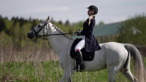 professional woman rider is sitting in saddle on white horse and riding in field at summer