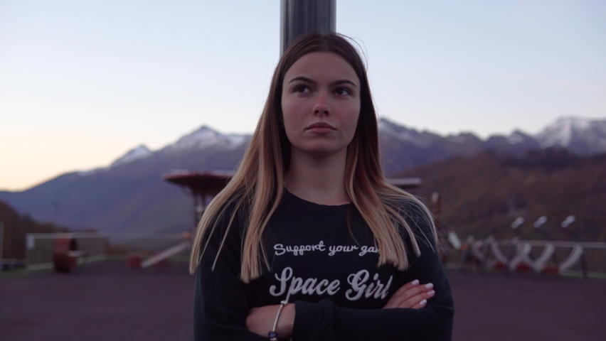 A girl with a cool look is standing on a sports field in the mountains leaning on a pole. She looks away. The camera moves backwards