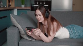 Young woman in headphones gamer using smartphone and play in online video game. She lying in couch playing online games in modern apartment background.