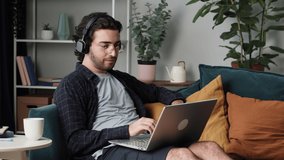 Young smiling man in glasses and headphones, speaking with friends or relatives, using online video call on laptop computer, drinking coffee and sitting on sofa. Happy guy talking using web camera. 