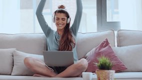 Video of motivated young woman dancing and listening to music with her laptop while sitting on sofa at home.