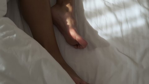 bare feet of woman sleeping on white bed linen in bedroom or in hotel room, enjoying morning dream and moving legs, closeup view