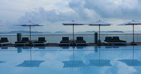 Static shot of blue swimming pool in foreground reflecting evening clouds, white beach umbrellas and lounge chairs. A dramatic oceanfront background with karst mountains in Phuket Thailand.