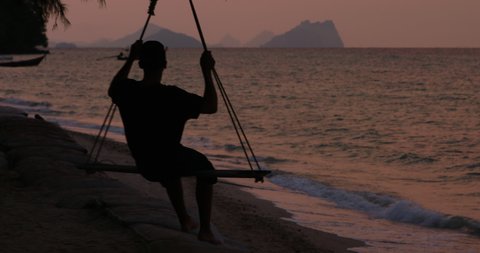Static shot of silhouette of male tourist on a beach swing during pink sunset on holiday in tropical paradise Thailand. Waves lap on the beach, longtail boats and mountains fill the background