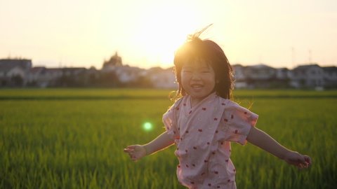Happy Asian little girl running in the sunset background with smile. Slow motion. Toddler kid in traditional Japanese summer clothing. Rice paddy fields, residential area. Healthy growth of children