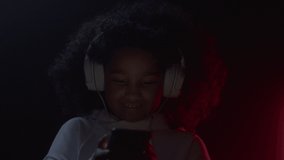 Portrait of little happy afro american girl listening music wearing headphones, using social media on smart phone in colorful neon light closeup indoors. Modern kid lifestyle, funny technology relax