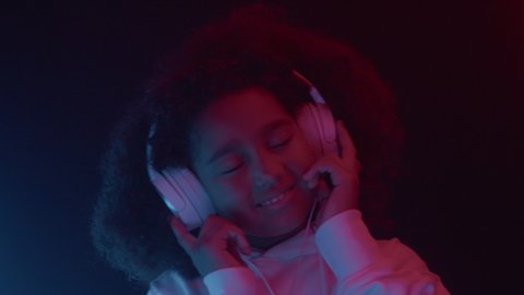 Portrait of happy african american baby girl listening music wearing headphones, dancing in colorful neon light indoors close up. Enjoy cool melody, funny leisure, modern relax using audio technology