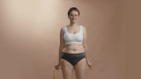 Chubby body positive female in white bra and black briefs measures her waist with measure tape and makes victory dance. Studio beige background still cowboy shot video.