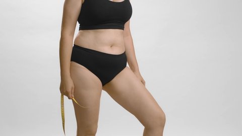 Full figured lady in black underwear wants to know her thighs volume using measure tape. White background still shot high quality studio video. Anonymous cowboy shot.