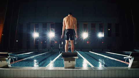 Muscular Male Swimmer Comes to Starting Block to Jump and Dives into Swimming Pool. Professional Man Athlete Ready to Win Championship. Cinematic Light, Slow Motion, Flare Light, Back View Wide Shot
