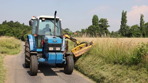 Much Hadham, Hertfordshire. UK. July 20th 2021. Man in a tractor using a flail to mow grass verges in a country road.