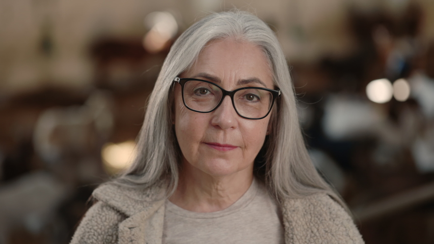 Portrait of grey haired charming woman in eyeglasses and casual wear posing at goat farm. Mature female farmer growing domestic animals for organic milk and meat.