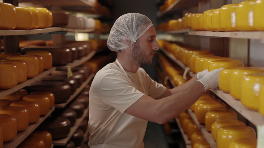 Young caucasian man in uniform and gloves checking quality of cheese heads that aging on shelves at storage house. Professional cheese maker keeping arms crossed, smiling and looking at camera.
