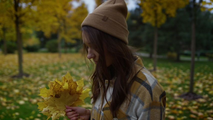 Side portrait view of smiling attractive girl walking with yellow maple leaves in hands with beautiful autumn background. Profile of stylish young woman wearing hat and plaid shirt outdoor.