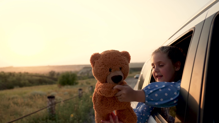Happy girl in car window with teddy bear.Happy family trip. Girl smiles at wind in car window. Family summer trip. Teddy bear in hand of child.Happy girl with family travel by car.Child smiles at wind