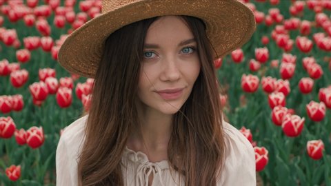Portrait of Gentle Lady in Straw Hat standing on the Field with Colorful Tulips. Looking into camera having Long Hair and Azure Eyes. Feeling Pleasure with Nature. Pretty Appearance of Girl. Romantic.