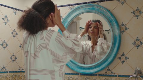 20s African American woman with pony tail admiring with her reflection in bathroom mirror at home. Woman likes herself during morning routine in bathroom.