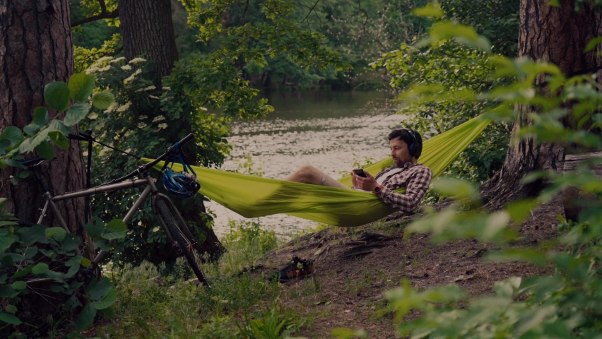Young caucasian man resting in hammock, listening to music on headphones and using smartphone afterwards on bicycle in forest near the lake. Audio healing. Idyllic place. Travel, camping in nature - Powered by Shutterstock - Get 15% off with code: PIKWIZARD15