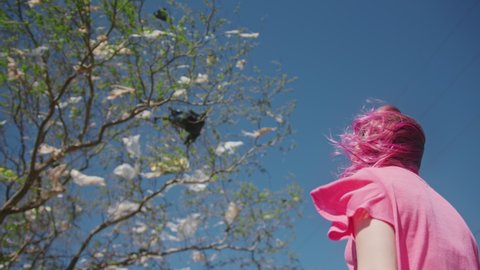 Back view of hipster woman with bright pink hairstyle staring horrified at green tree covered by the plastic bags caught up on branches. Degrading nature landscape against blue sky, slow motion 4K