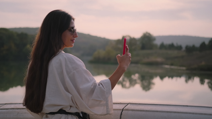 Young brunette is delighted with peaceful river landscape and taking photos with smartphone from the bridge