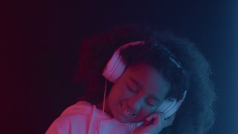 Portrait of smiling cheerful african american child girl listening cool music wearing headphones, having fun in colorful neon light indoors close up. Happy modern relax, audio enjoyment using device