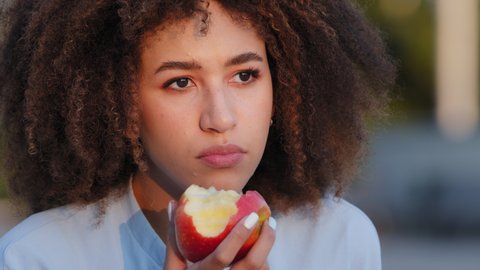 Extreme close-up portrait outdoors african american woman with curly hair afro hairstyle pensive lady female eating biting red fresh delicious natural apple enjoying eat fruit sad food diet chewing