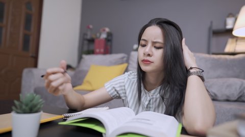 Attractive asian black hair female student having a hard time understanding on her homework, sitting upset unhappy on living room floor, scratching head while contemplating on solving problems, 