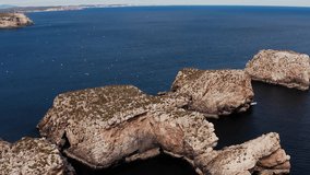 Aerial view of brown rocky islands in the open blue water. Drone flight over calm Atlantic ocean in Portugal with a few clouds and seagulls flying around. On top of the cliffs is some green grass.