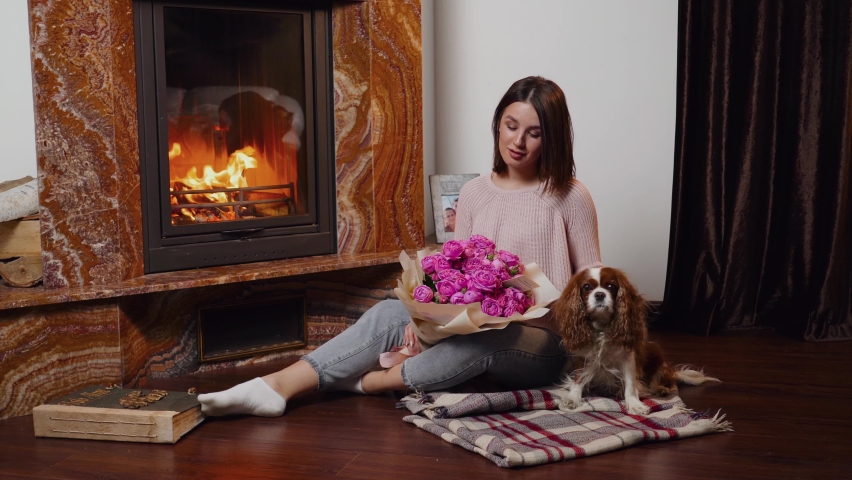 A young woman holds a beautiful bouquet of roses, sitting on a blanket next to a dog near a burning fireplace.