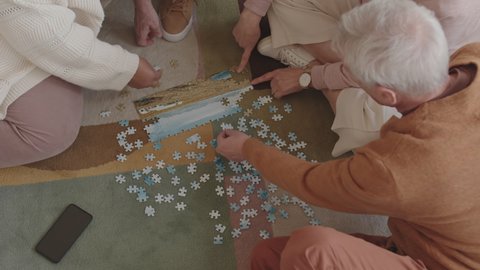 Top view of group of unrecognizable people sitting on carpet on floor, doing puzzle