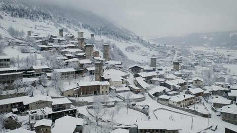 This is an aerial drone camera shot of Mestia village in Svaneti region, Georgia, Caucasus mountains. Shot taken on a cloudy winter day. Svan defence houses – towers covered in snow