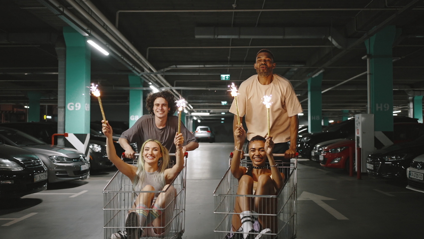 Group of happy diverse friends riding on shopping carts at underground parking, ladies holding burning fireworks, slow motion Royalty-Free Stock Footage #1076308541