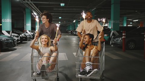 Group of happy diverse friends riding on shopping carts at underground parking, ladies holding burning fireworks, slow motion
