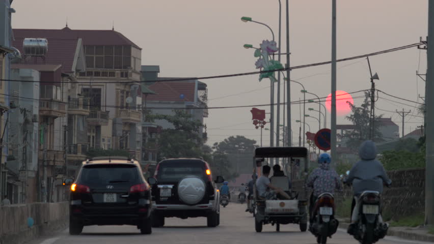 Large orange red sun sets slowly in background while cars and scooters move towards the sunset - returning home after a full day of work against an urban backdrop of Hanoi, Vietnam