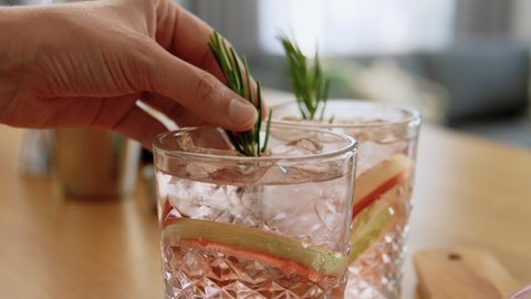 drinks and people concept - young woman making rhubarb cocktail and decorating it with sprig of rosemary at home kitchen