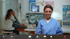 Dentist nurse talking with remote patients about hygiene looking at camera in dental center while doctor is working. Stomatologist assistant speaking on webcam, footage with software overlay concept - Powered by Shutterstock - Get 15% off with code: PIKWIZARD15