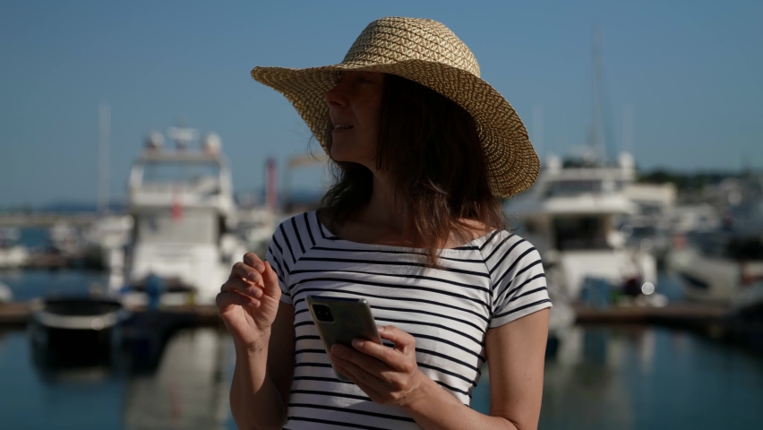 an adult woman in a big hat is standing with a mobile phone in her hand on a walk to the yacht port
