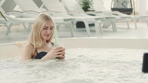 Medium shot of charming blonde woman resting in jacuzzi with soft drink. Spending vacations at the resort.